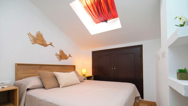 Quarto com cabeceira de madeira, cama com roupa de cama bege, mesa de cabeceira com abajur e guarda-roupa.