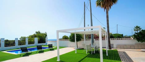 Un área de piscina en la azotea con césped artificial, una pérgola y una palmera.