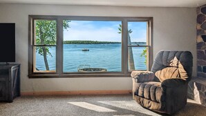 A room with a large window overlooking a lake, a stone wall, and a cushioned armchair. - The Cove on Lake Carlos (Carlos Township)