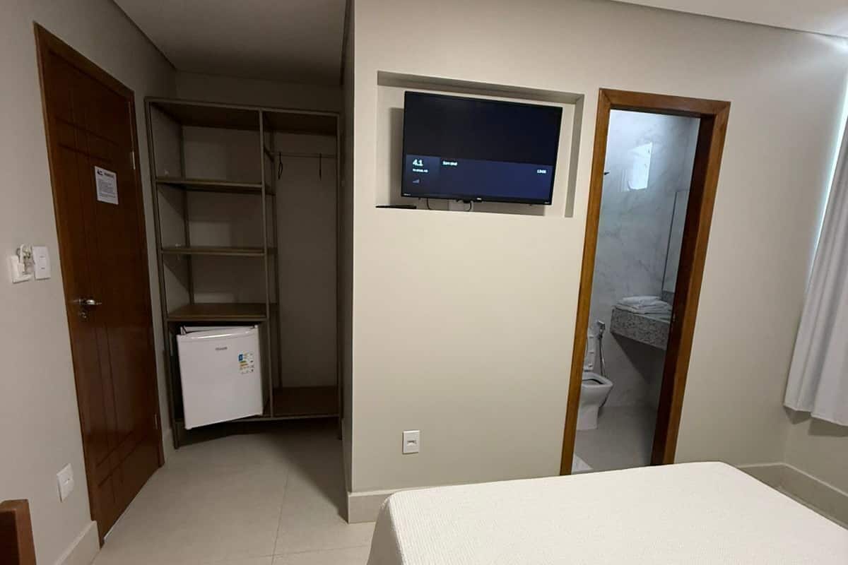 A bedroom with a bed, a TV mounted on the wall, a wardrobe, and a small refrigerator.