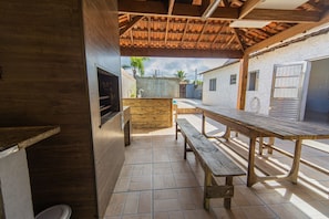 An outdoor patio with a wooden table, benches, a grill, and a view of a residential area. - House with Pool 5 Minutes from Gaivotas Beach (Itanhaém)