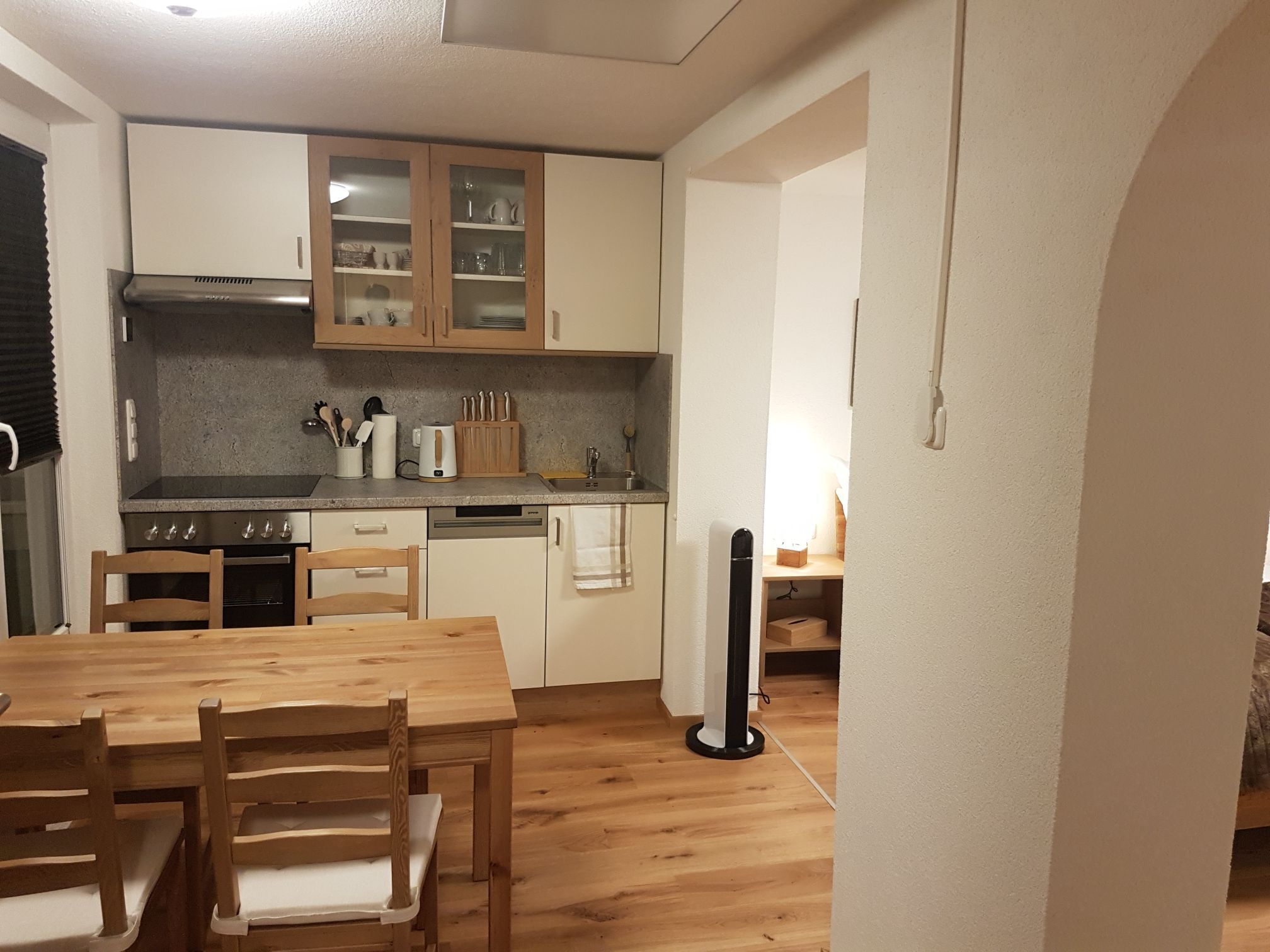 Une cuisine de taille réduite, dotée de meubles en bois, d’une table à manger, de chaises et d’un ventilateur sur pied.