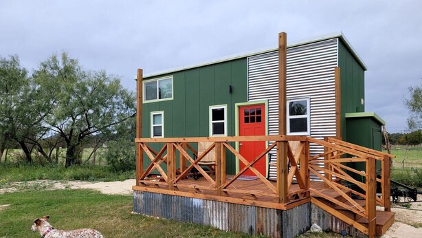 A green building with a red door, a wooden deck, and a dog in the foreground. - Gorgeous Tiny House with Spectacular Countryside Vistas in Camp Wood, Texas (Camp Wood)