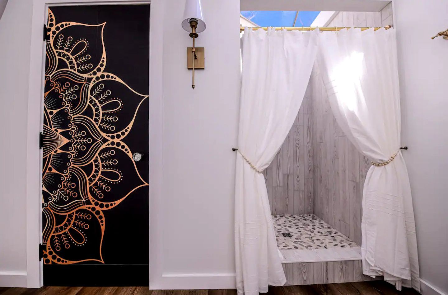 A room with a black door featuring a gold paisley pattern, a white canopy bed with a patterned bedspread, and a wall-mounted light fixture.