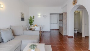 Una sala de estar con un sofá, una mesa de comedor con sillas, una planta pequeña y un reloj en la pared.