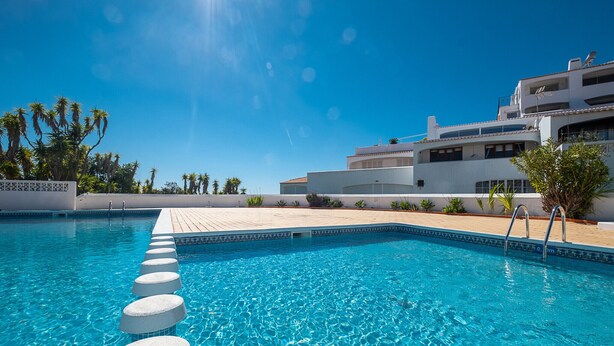 Una piscina con agua azul cristalina, un edificio blanco con balcones y palmeras al fondo.