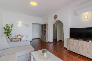 Una sala moderna con un sofá, una mesa de comedor con sillas, un televisor sobre un mueble bajo y un pasillo que conduce a otra habitación.