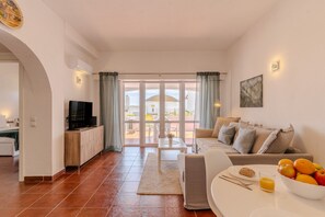 Una sala moderna con un sofá, una mesa de centro, un televisor y un comedor con un tazón de fruta y un vaso de jugo.