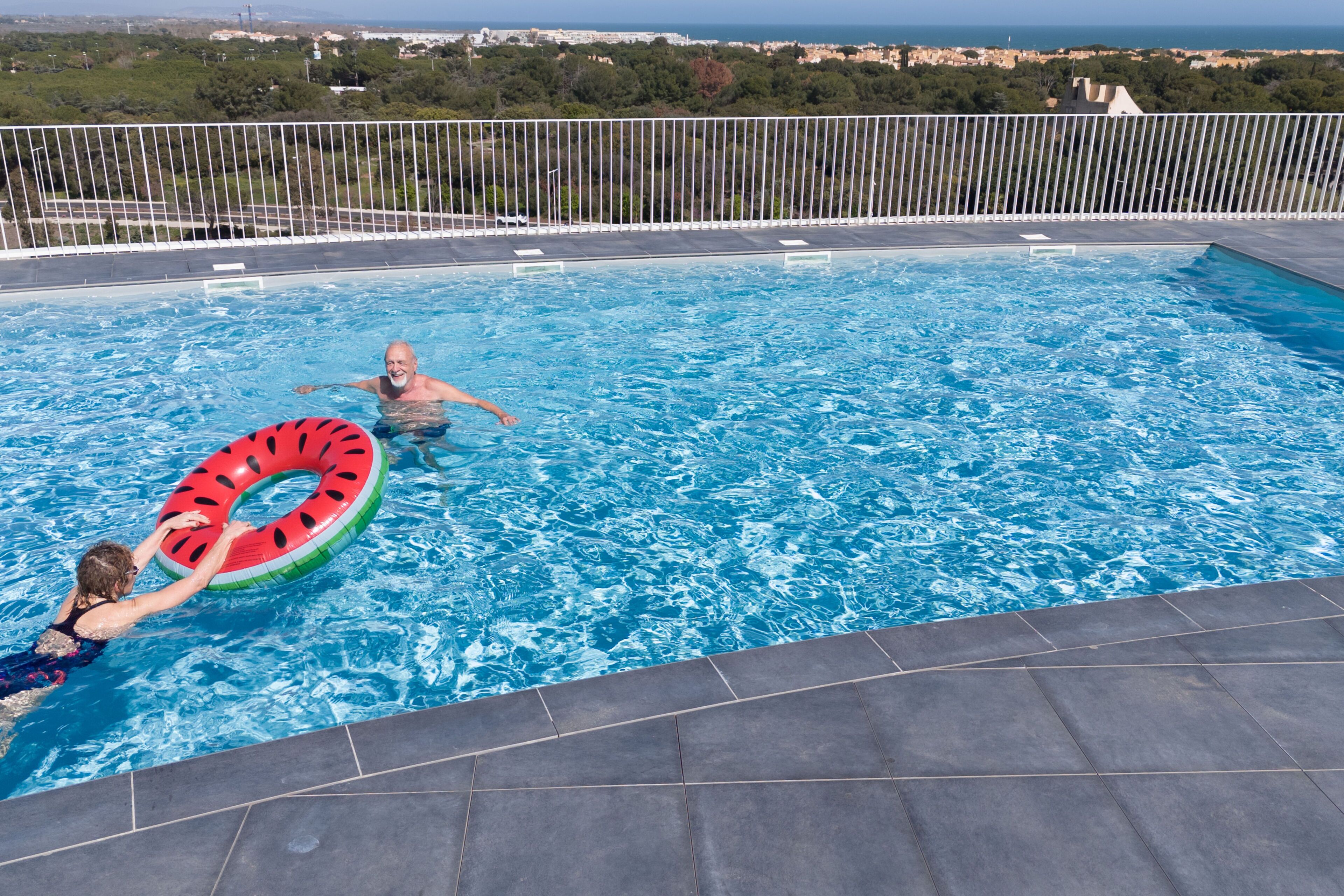 Ein Swimmingpool mit klarem, blauem Wasser, einer gefliesten Liegefläche und Blick auf das Meer und die grüne Landschaft im Hintergrund.