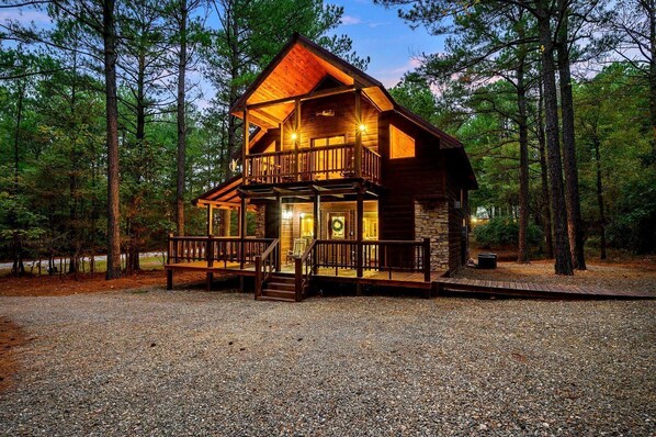 A two-story log cabin with a balcony, surrounded by a gravel driveway and forest. - Silver Spur (Broken Bow)