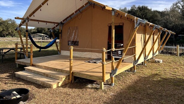 A temporary structure with a raised platform, a hammock, and a sign welcoming guests. - Deluxe Safari Tent with Fire Pit, Hammocks & BBQ in Basinger, Florida (Okeechobee)