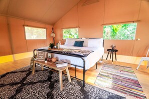 Une chambre spacieuse comprenant un lit, des tables de chevet, un banc et une vue sur la verdure grâce à de grandes fenêtres.