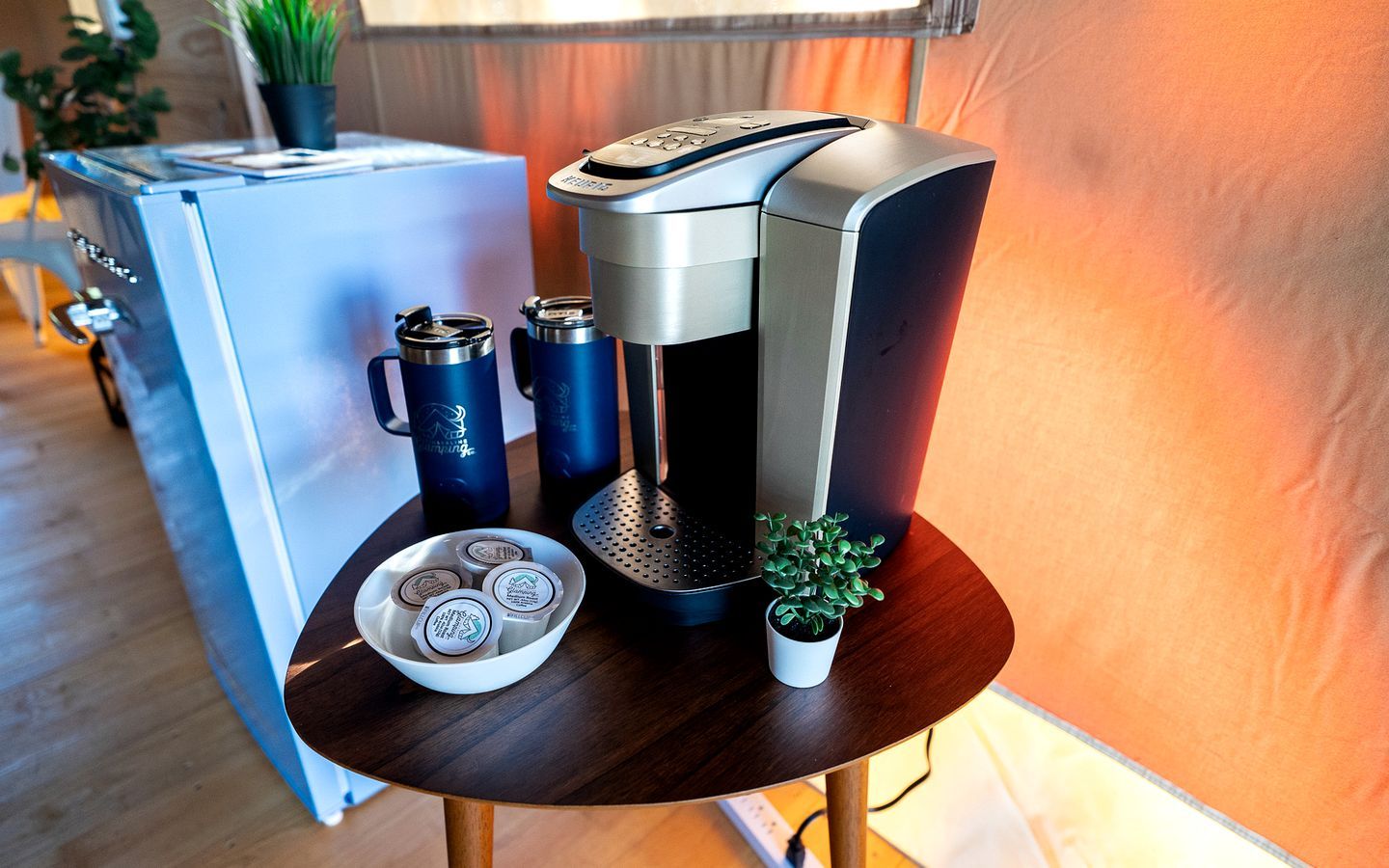 A coffee machine on a wooden table with a bowl of coffee pods, a thermos, and a potted plant.