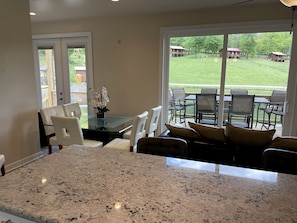 Une cuisine dotée de plans de travail en granit, un coin repas avec des chaises blanches et une grande porte vitrée donnant sur une terrasse aménagée avec du mobilier d’extérieur.