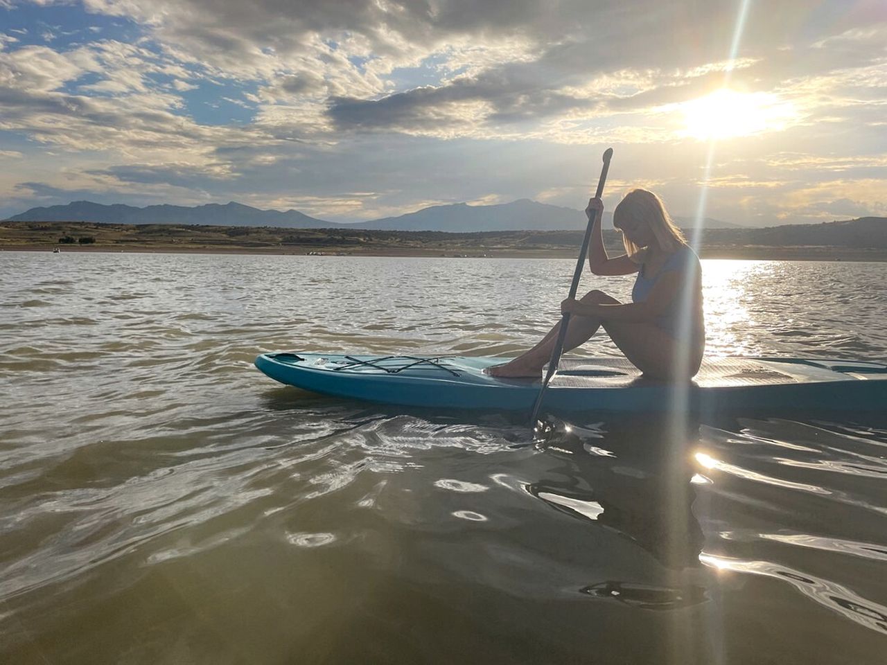 Eine Person steht auf einem Paddleboard auf ruhigem Wasser, mit blauem Himmel und Bergen im Hintergrund.