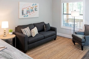 Ein modernes Wohnzimmer mit einem dunkelgrauen Sofa, einem runden Beistelltisch mit Lampe, einem kleinen Topfpflanzen und einem Fenster mit Jalousien.