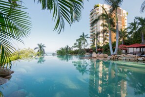 A clear view of a swimming pool surrounded by palm trees and a multi-story building with balconies. - 5-Diamond Resort ◆ Garza Blanca PV 3BR ◆ Save 45% vs. Garza Blanca Direct (Puerto Vallarta)