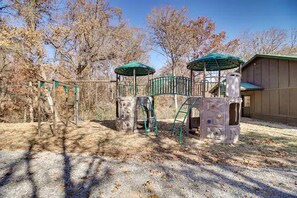 Un parque infantil con estructuras para escalar y una zona cubierta, rodeado de árboles y un edificio.