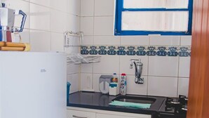 A compact kitchen with white cabinets, a black countertop, a microwave, and a window with blue blinds. - Apto Climatizado com Wi-fi no Centro de Bel m (Belém)