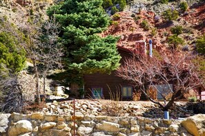 A cabin with a stone wall in front, surrounded by trees and a rocky hillside.