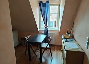 A small, triangular room with a wooden table and chairs, a radiator, a window with curtains, and a desk with a basket and papers.