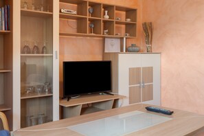 A modern living room with a flat-screen TV, wooden shelving with decorative items, and a glass-top coffee table.