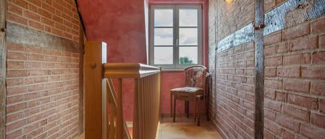 A narrow hallway with a wooden railing, a chair, a window, and a brick wall.