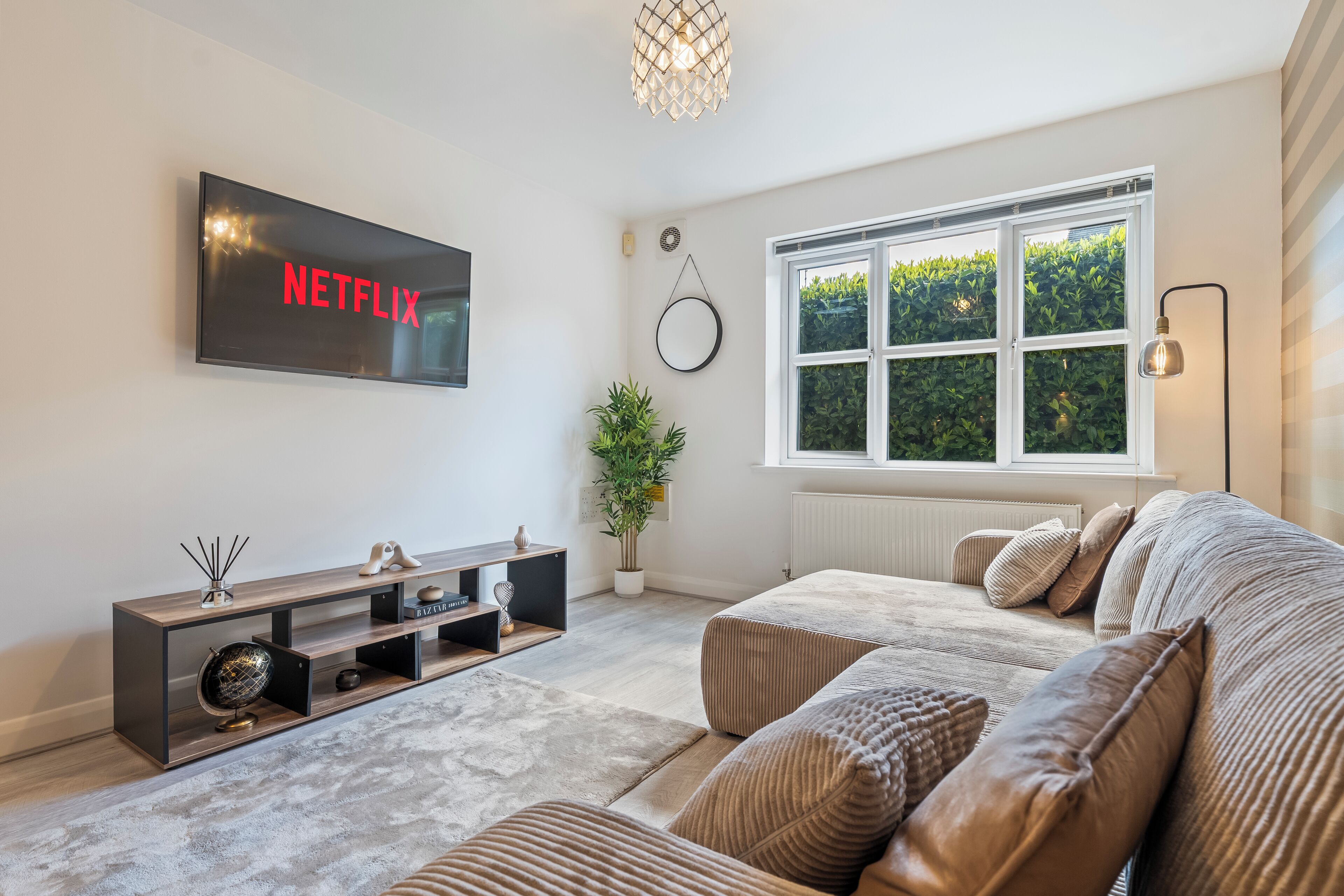 A modern living room with a flat-screen TV, a sofa, a coffee table, a potted plant, and a large window.
