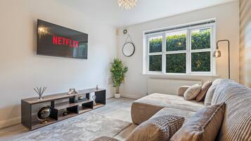 A modern living room with a flat-screen TV, a sofa, a coffee table, a potted plant, and a large window.