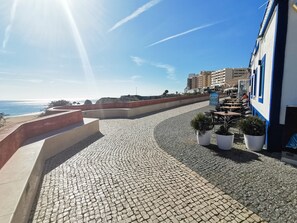 Eine Kopfsteinpflasterpromenade mit Außenbestuhlung, mit Blick auf eine Küstenlandschaft und Gebäude im Hintergrund.