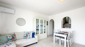A modern living room with a sofa, dining table, chairs, a cabinet with glass doors, and an air conditioning unit. - Apartment Praia Rocha | Sea | Concorde Infinity (Portimão)