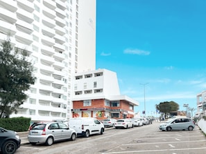 Ein Parkplatz mit mehreren Autos, einem Supermarkt und einem hohen, weißen Gebäude mit Balkonen.