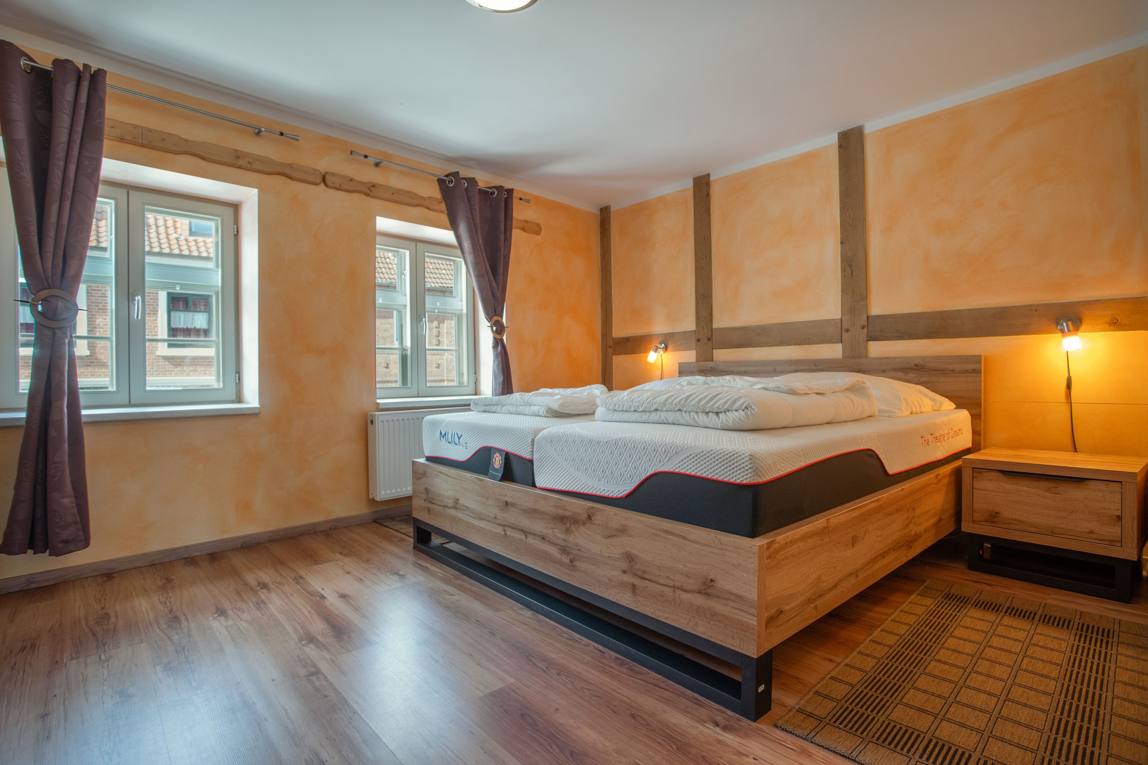 Une chambre à coucher avec un lit en bois, une table de chevet, une fenêtre avec des rideaux et un radiateur.