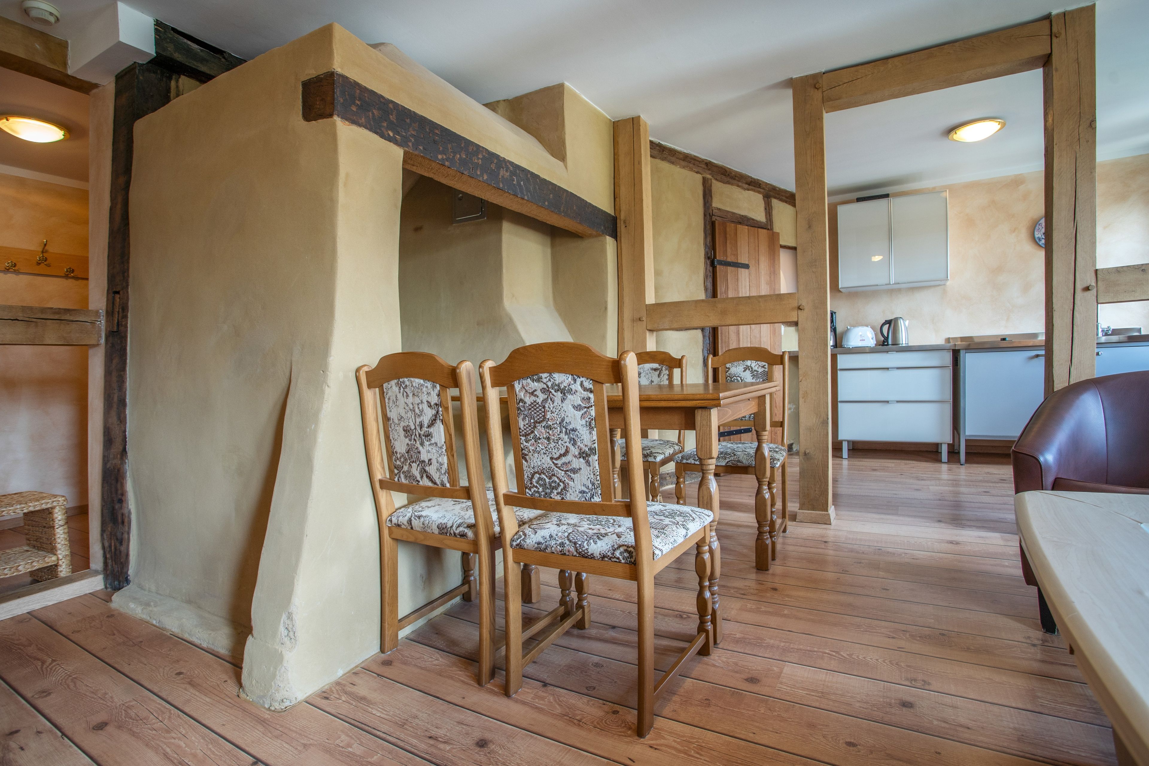 A dining area with wooden chairs and a table, a kitchen in the background, and a wooden floor.