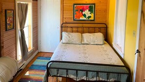 A bedroom with a wooden ceiling, a bed with a white blanket, a colorful striped rug, and a painting on the wall.
