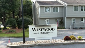 Schild für Westwood at Bethany, ein Gebäude mit grauem Mauerwerk, einer Auffahrt und einer bepflanzten Grünfläche mit Steinen und Pflanzen.