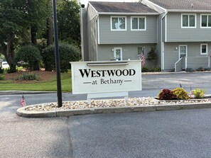 A sign for Westwood at Bethany, a building with a gray exterior, a driveway, and a landscaped area with rocks and plants. - Bethany Coastal Retreat, Fully Updated Home, Pool Access & under 1 mile to Beach (Bethany Beach)