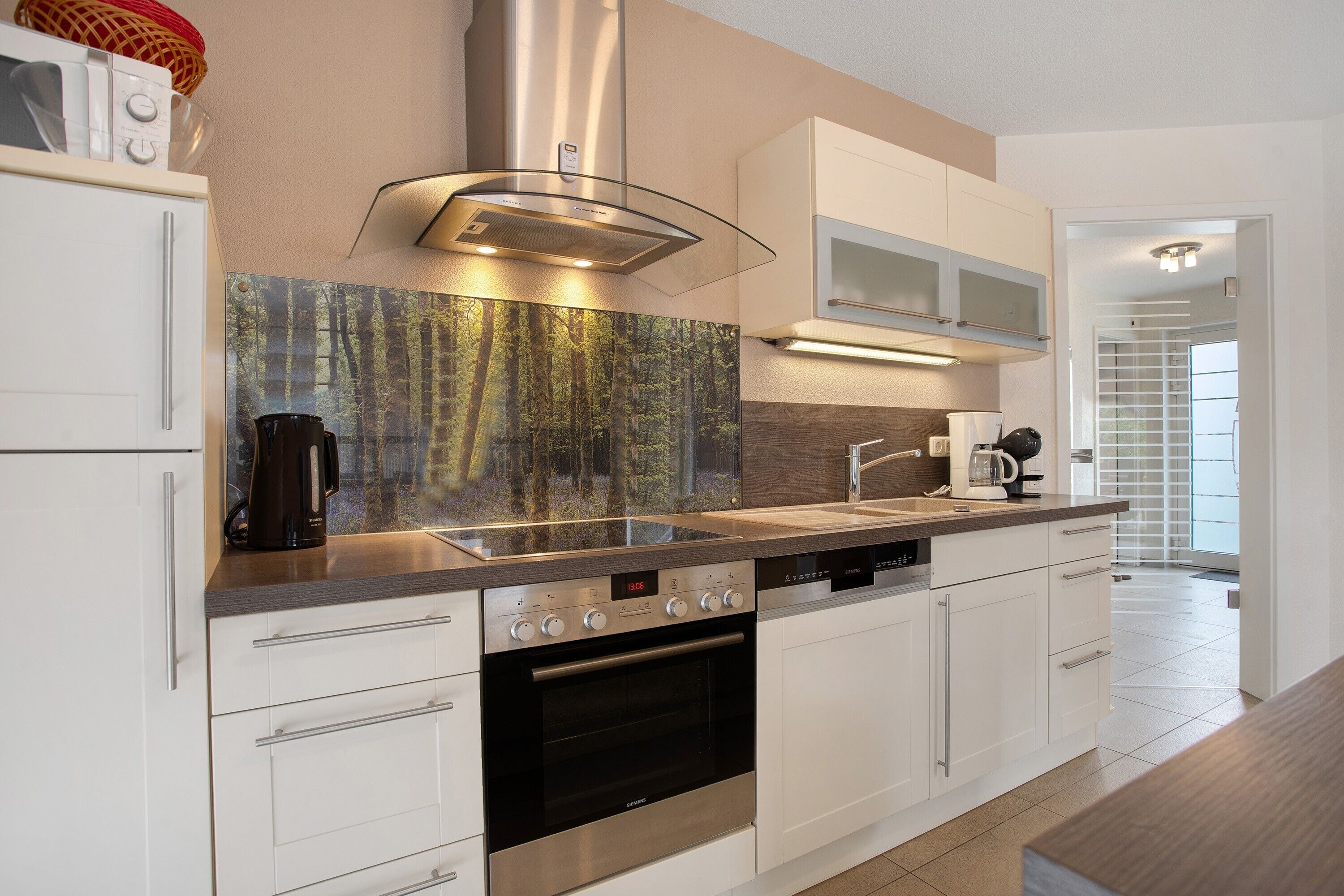 A modern kitchen with white cabinetry, a stainless steel range hood, a built-in oven, and a large window with a forest view.