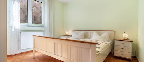 Quarto com cama, mesas de cabeceira, janela com cortinas e radiador.