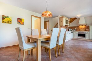 A dining area with a wooden table and chairs, a hanging light fixture, and kitchen cabinets in the background.