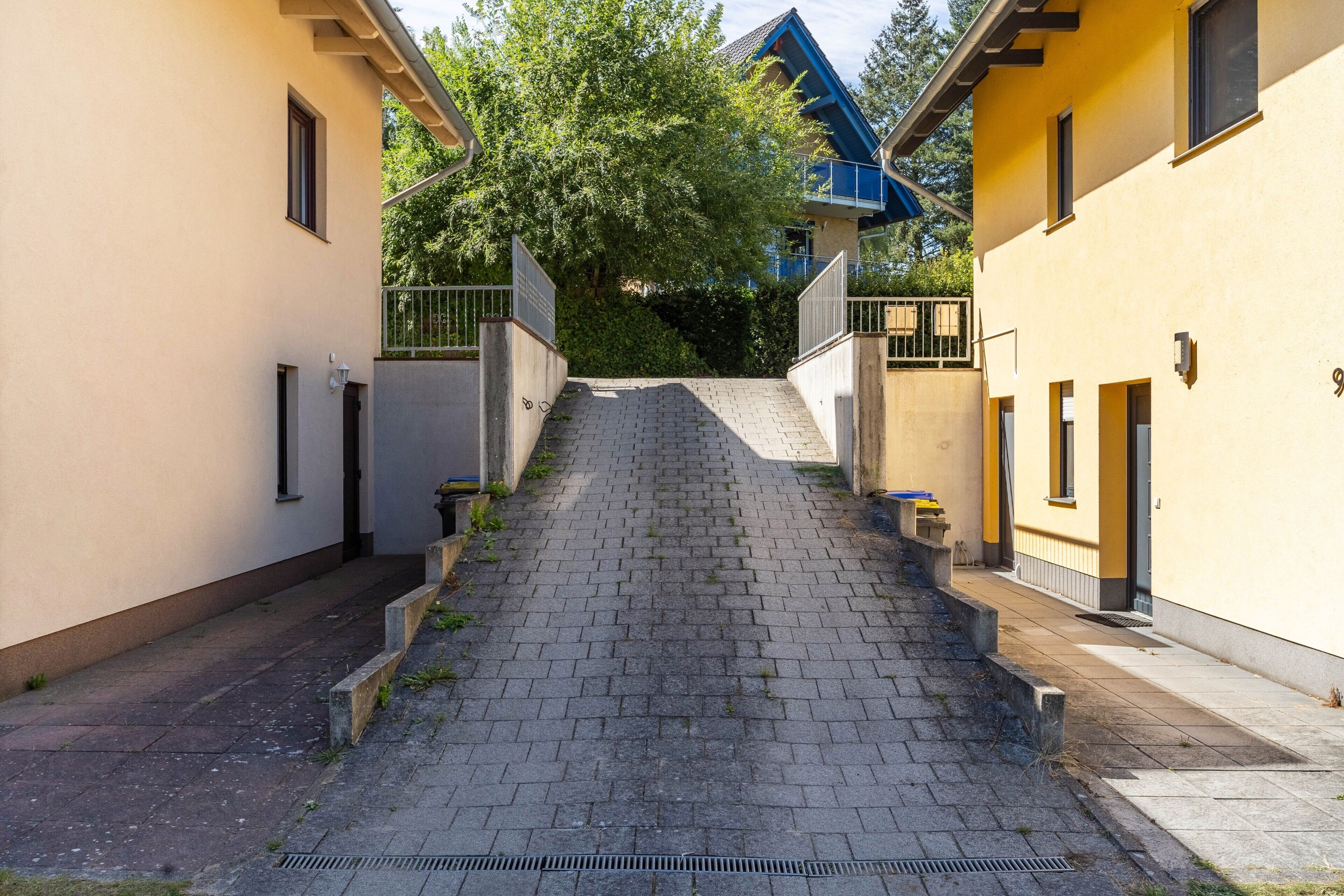 Uma estreita passagem pavimentada leva a um edifício com telhado azul, ladeado por dois edifícios com paredes bege e janelas pequenas.