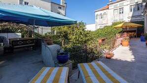 Een terras buiten met een blauw dakzeil, tafels met gele strepen en potplanten.