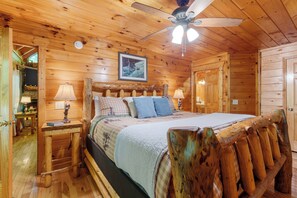 A log cabin bedroom with a bed, wooden headboard, two bedside tables with lamps, a ceiling fan, and a framed picture on the wall. - Log Cabin near Pigeon Forge | Hot Tub, Fireplace (Sevierville)