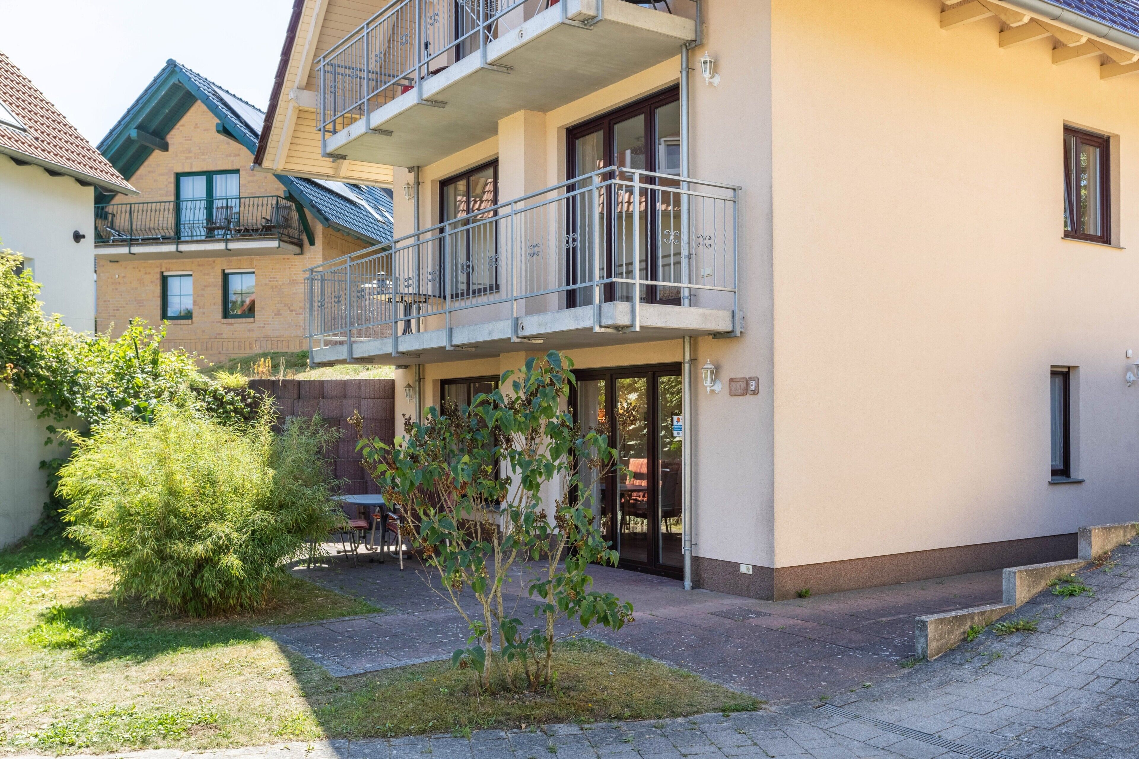 Un immeuble résidentiel à plusieurs étages, doté de balcons, d’une cour pavée et d’un petit jardin.