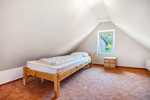 Una habitación pequeña con una cama individual, techo inclinado, ventana y mesita de noche de madera.