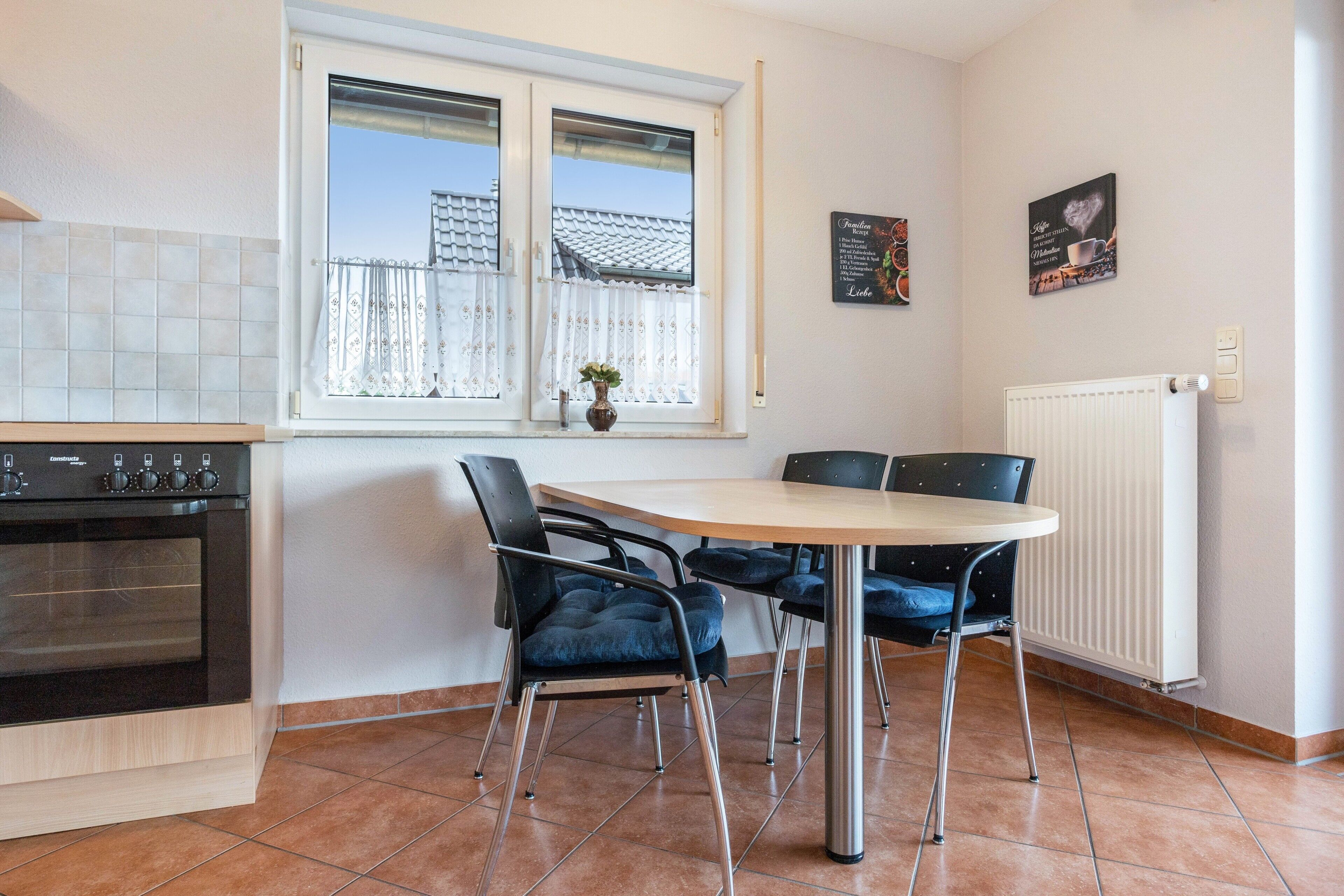 Een keuken met een fornuis, een eettafel met stoelen, een raam met gordijnen en een radiator.