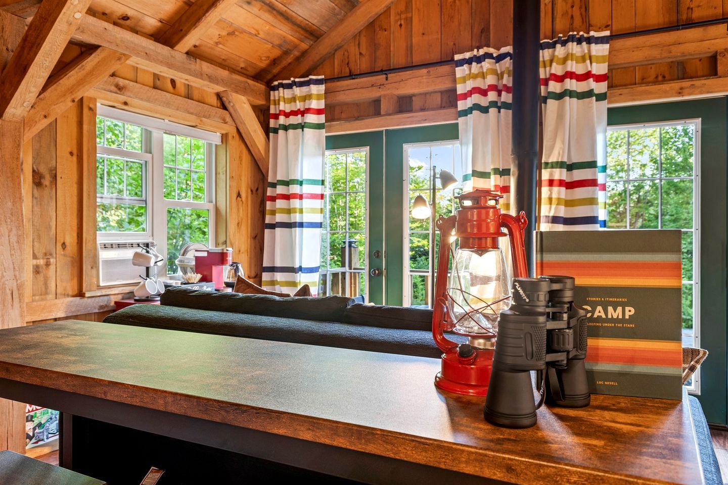 A cabin interior with a wooden ceiling, a wooden bench, a table with a lantern, a coffee maker, and a book titled 'CAMP'.