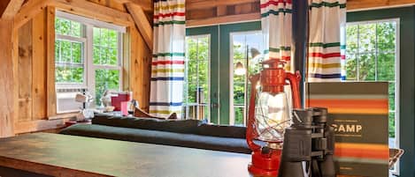 A cabin interior with a wooden ceiling, a wooden bench, a table with a lantern, a coffee maker, and a book titled 'CAMP'.