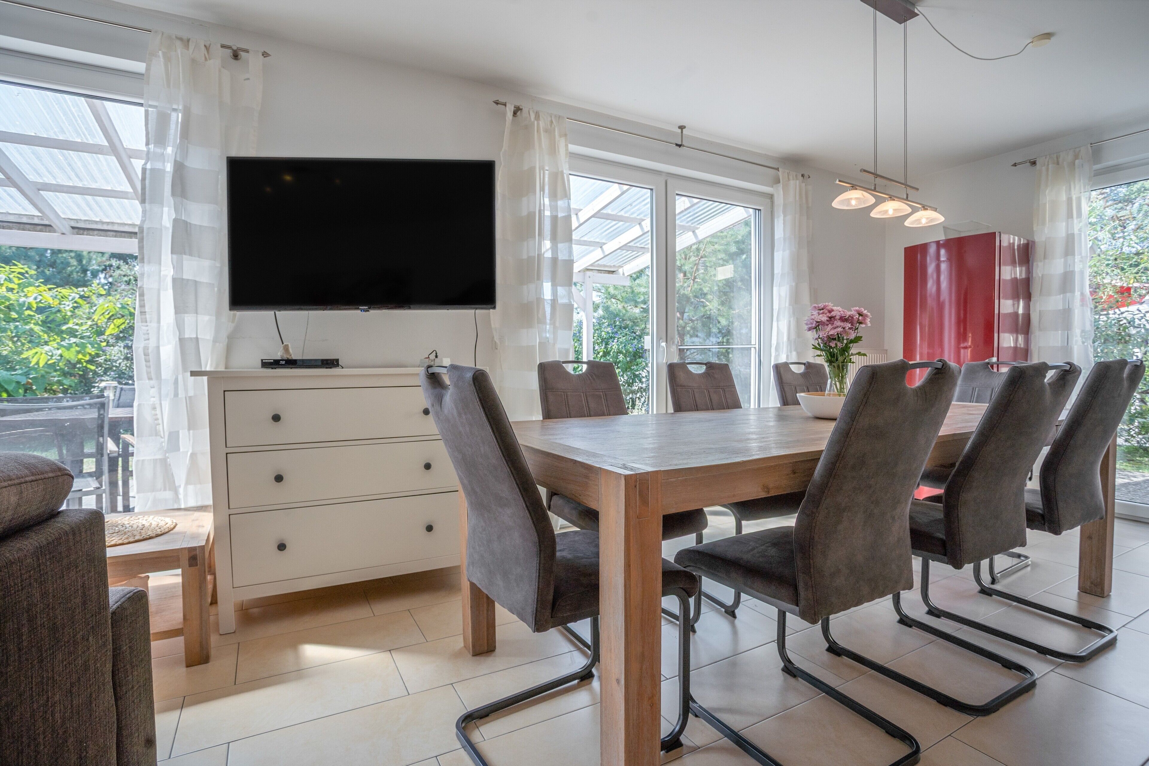 Un espace repas moderne avec une table en bois, six chaises, un téléviseur à écran plat et une grande fenêtre avec des rideaux.