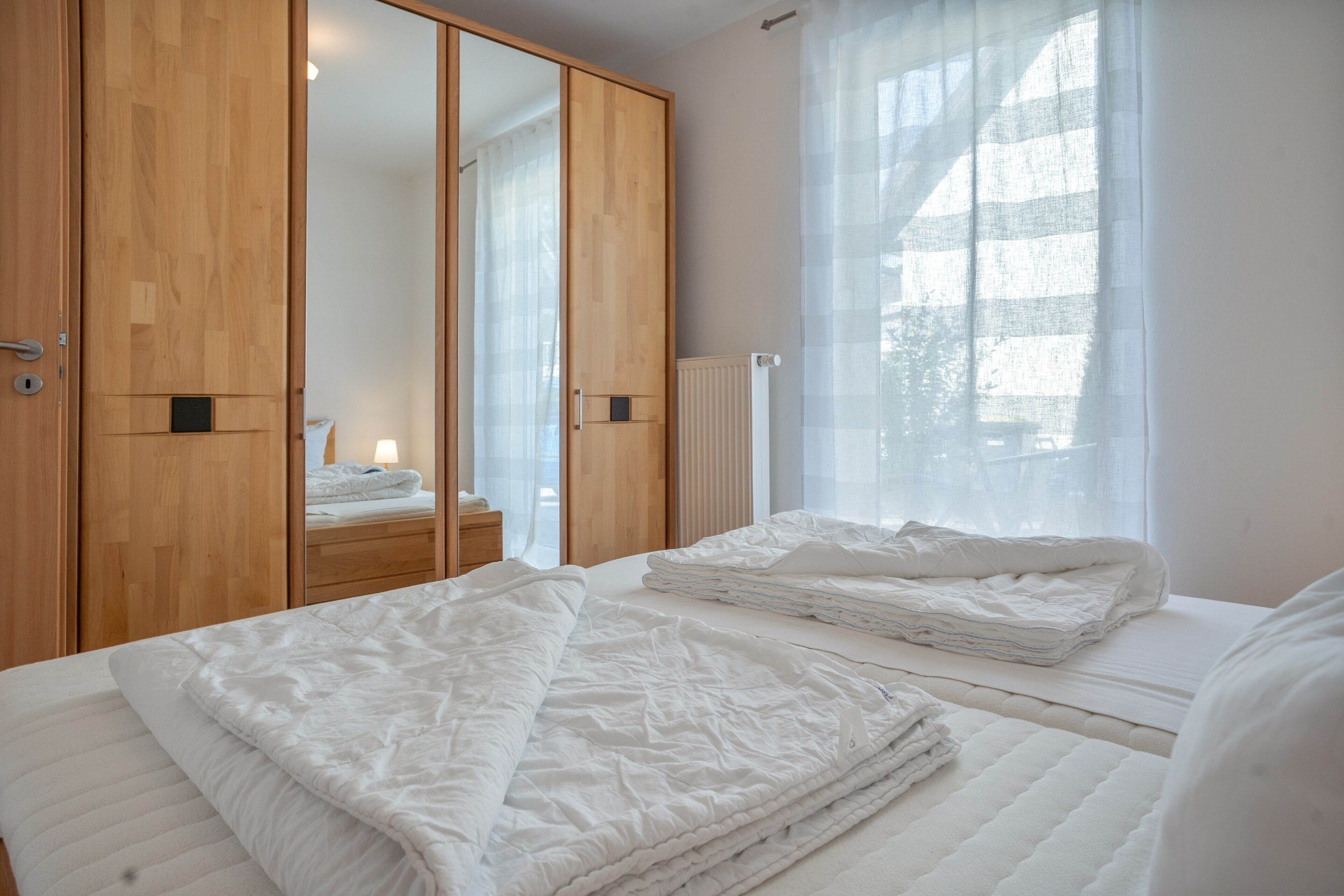 A hotel room with a large wardrobe, a bed with white bedding, and a window with sheer curtains.